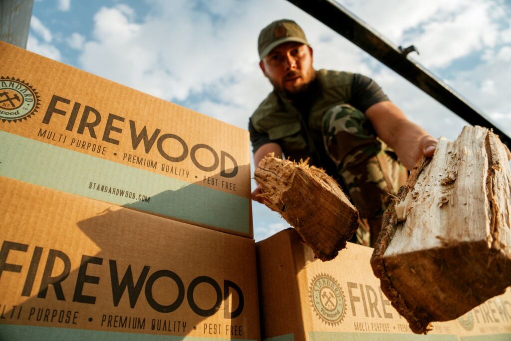 Worker holding pieces of split wood beside stacked Standard Firewood boxes
