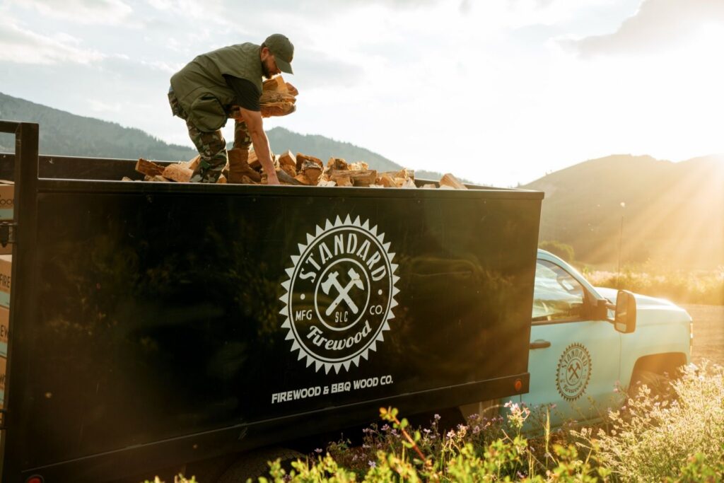 Worker handling split firewood in a black Standard Firewood trailer with the company logo, hitched to a light blue pickup near Park City, Utah